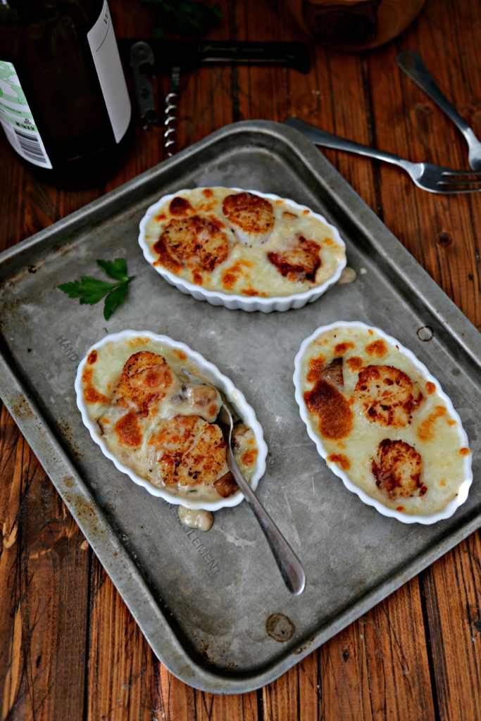 3 ramekins of Scallops Coquilles St. Jacques on a baking sheet. Bottle of wine, corkscrew, small forks and salt cellar in background.