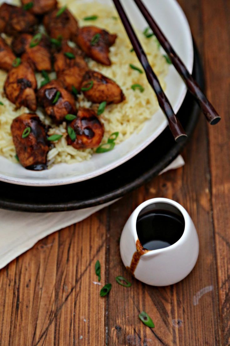 White bowl of bourbon chicken over sticky rice. Chopsticks and small jar of glaze to sides.