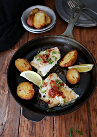 pieces of cod in a black cast iron skillet with potatoes and lemon wedges