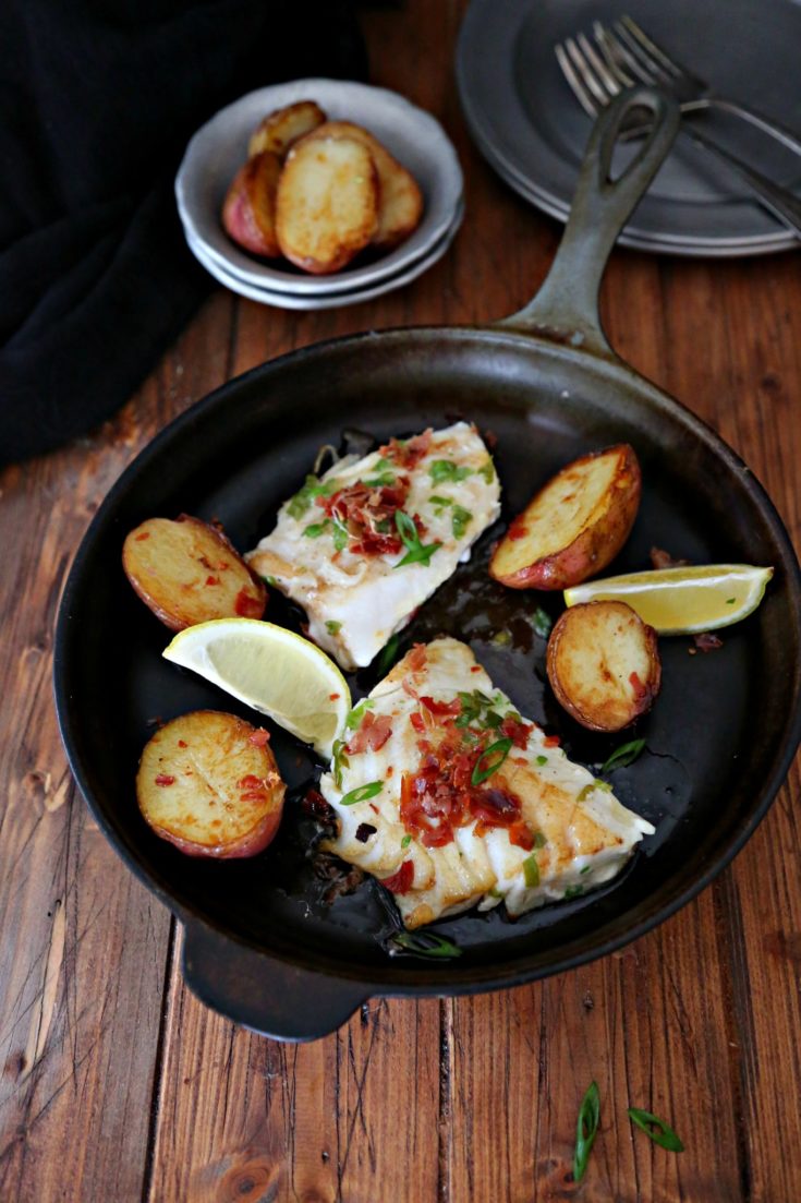 pieces of cod in a black cast iron skillet with potatoes and lemon wedges
