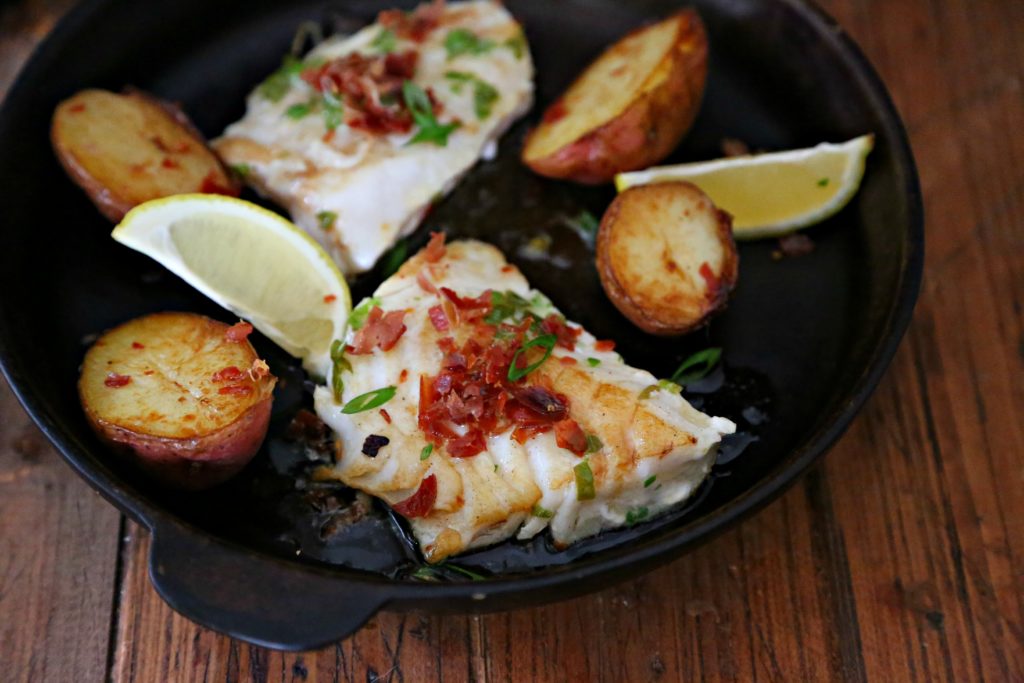 pices of cod in black cast iron skillet with potatoes and lemon wedges