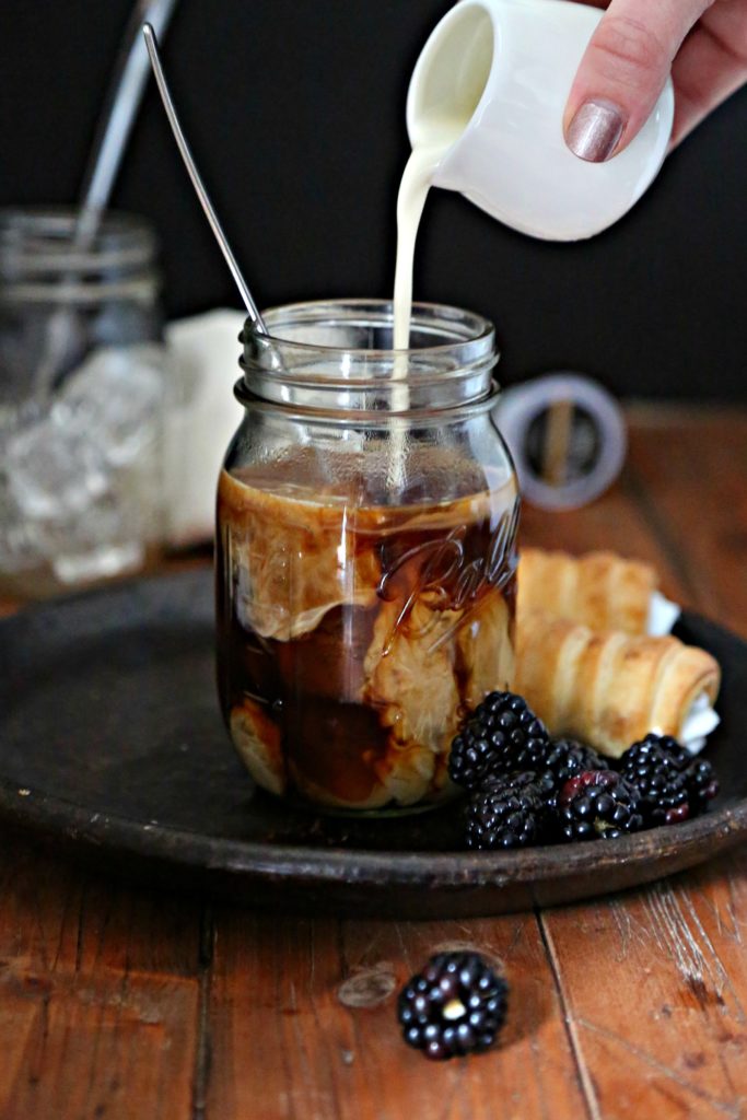 cream pouring into coffee over ice on dark plate with berries and pastry