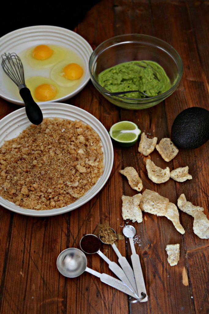 White bowl of crushed pork rinds. White bowl with eggs and whisk. Glass bowl with smashed avocado. Lime wedge, avocado, pork rinds and measuring spoons to right. 