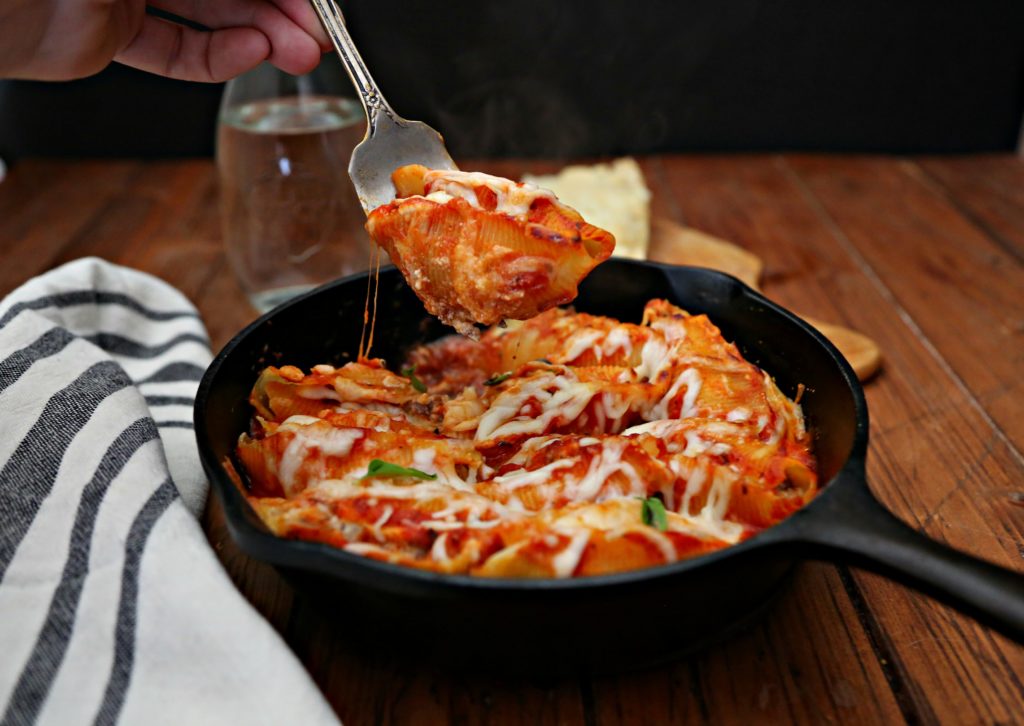 Bolognese Stuffed Shells in skillet, serving fork lifting one shell out. Carafe of water behind skillet. 