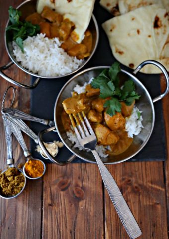 two bowls of Indian butter chicken with rice and naan bread. Measuring spoons filled with spices surrounding