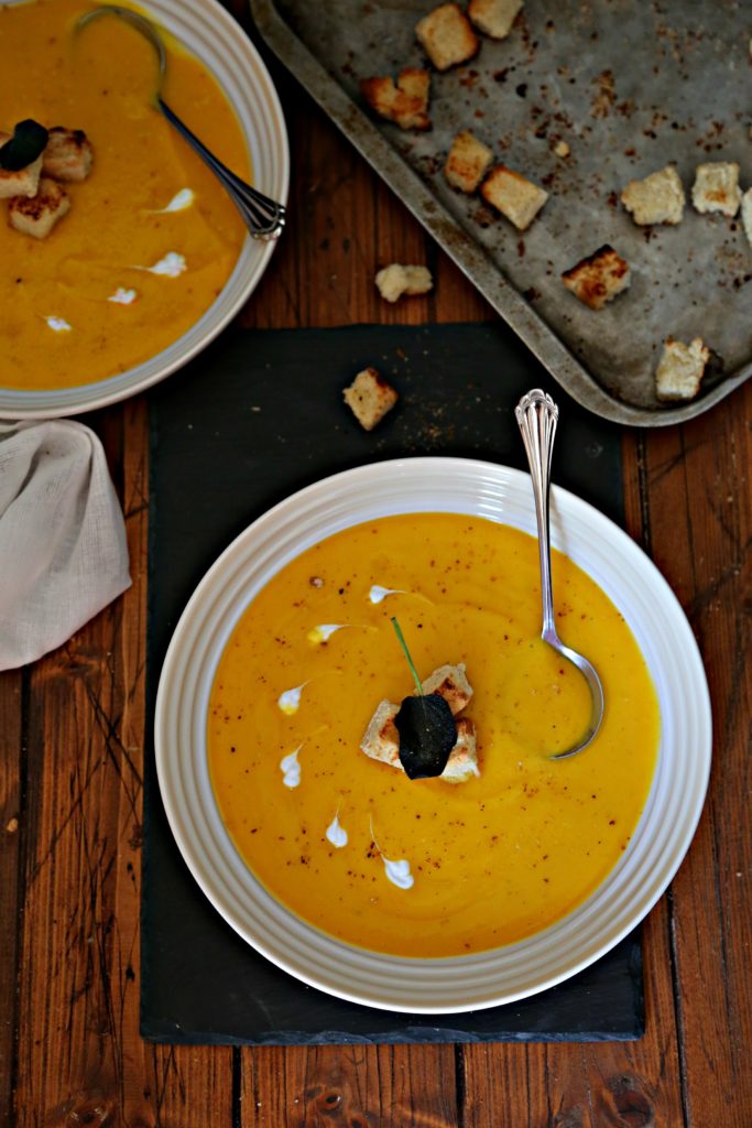 2 bowls of Roasted Butternut Squash Soup with spoons with a tray of croutons behind