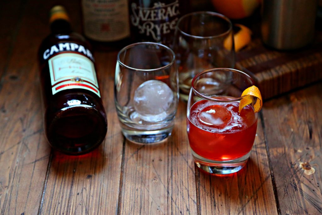 Boulevardier Cocktail with ice sphere and orange peel, extra glasses, bottle of campari and rye whiskey to side