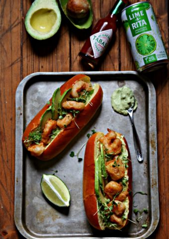 Spicy Shrimp Po Boy with Avocado Crema on baking sheet with lime, Tabasco bottle, avocads and lime a rita to side