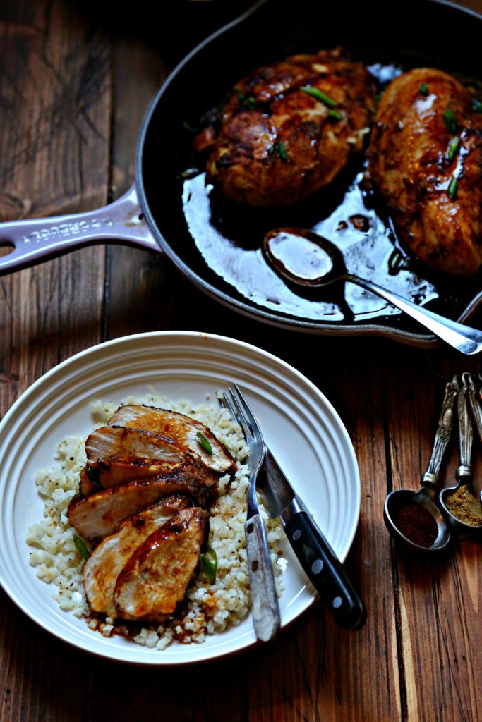 Spicy Skillet Chicken breasts with Pan Sauce in cast iron skillet with spoon. Bowl of sliced chicken over cauliflower rice with fork and knife. Measuring spoons with spices off to side.