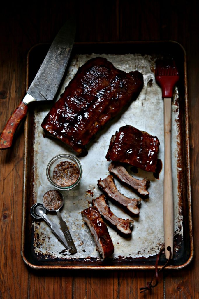 baking sheet with rack of bbq baby back ribs. Spice jar, measuring spoons, knife and red siicone brush.