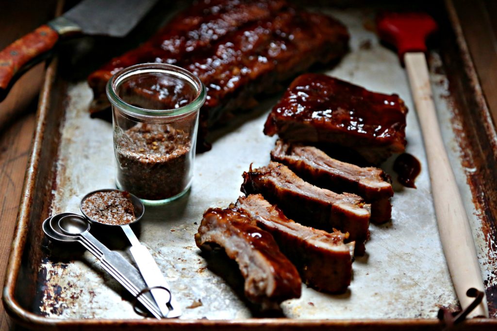 baking sheet with rack of baby back ribs, some sliced ribs, jar of spice rub, measuring spoons and red silicon brush.