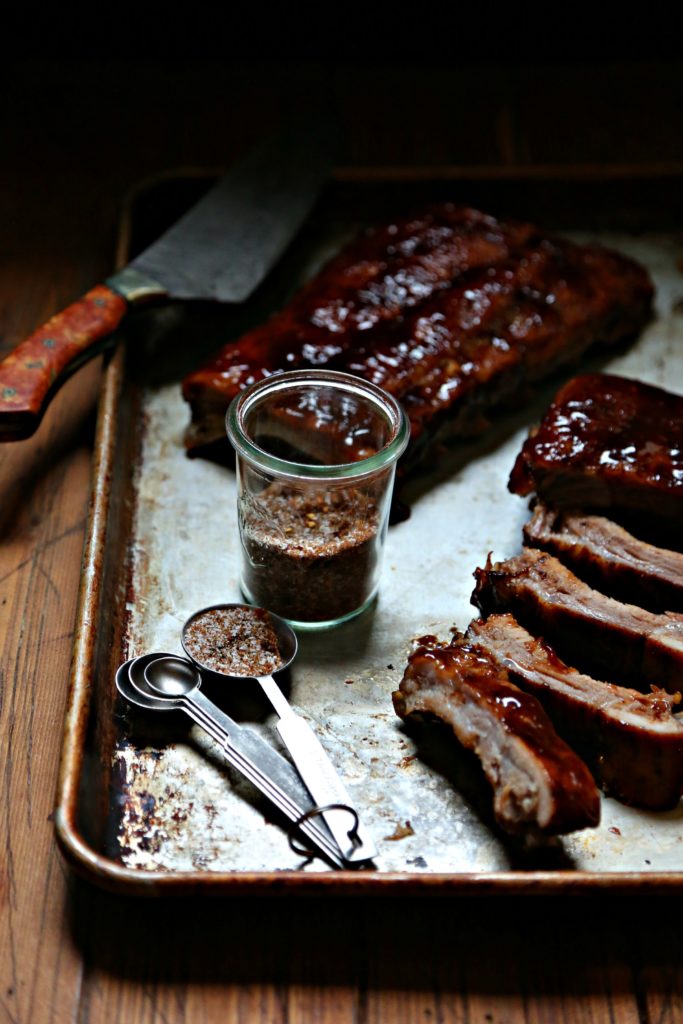 baking sheet with rack of ribs, sliced ribs, jar of spice rub, measuring spoons and knife.