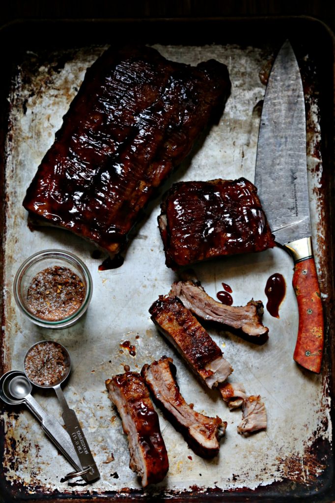 Sheet pan with rack of bbq baby back ribs. Jar of spice rub, measuring spoons and knife.