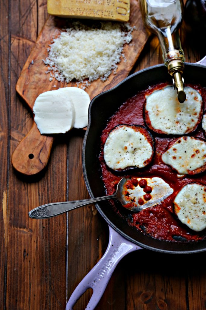 Skillet Eggplant Parmesan. Cutting board with parmesan and fresh mozzarella slices behind.Olive oil bottle to side. 