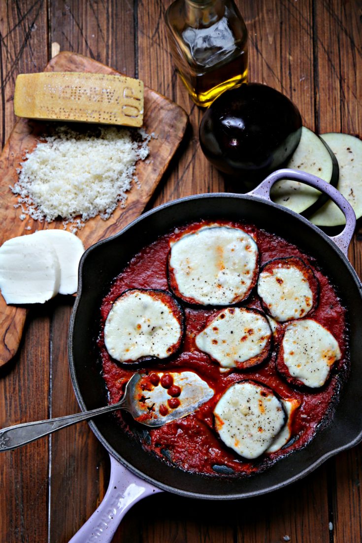 Skillet Eggplant Parmesan. Cutting board with Parmesan and mozzarella to side. Sliced eggplant behind.