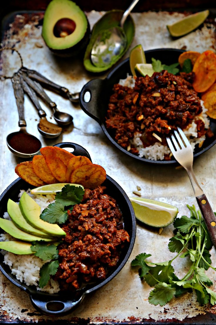 2 small cast iron skillets filled with rice, picadillo, avocado, cilantro, plaintain chips. Measuring spoons with spices behind.
