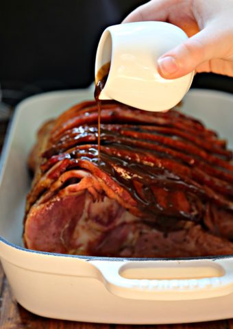 Balsamic Honey Dijon Glazed Spiral Ham in white roaster pan. Hand holding jar pouring glaze over ham.