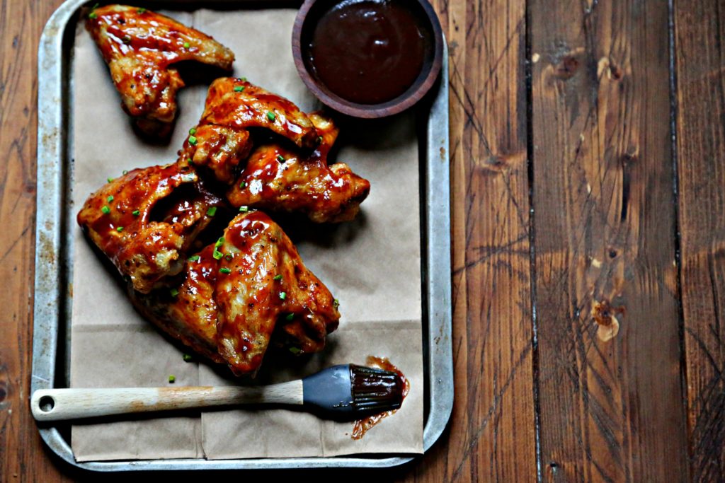 chicken wings on baking sheet with small bowl of bbq sauce and a silicone brush.