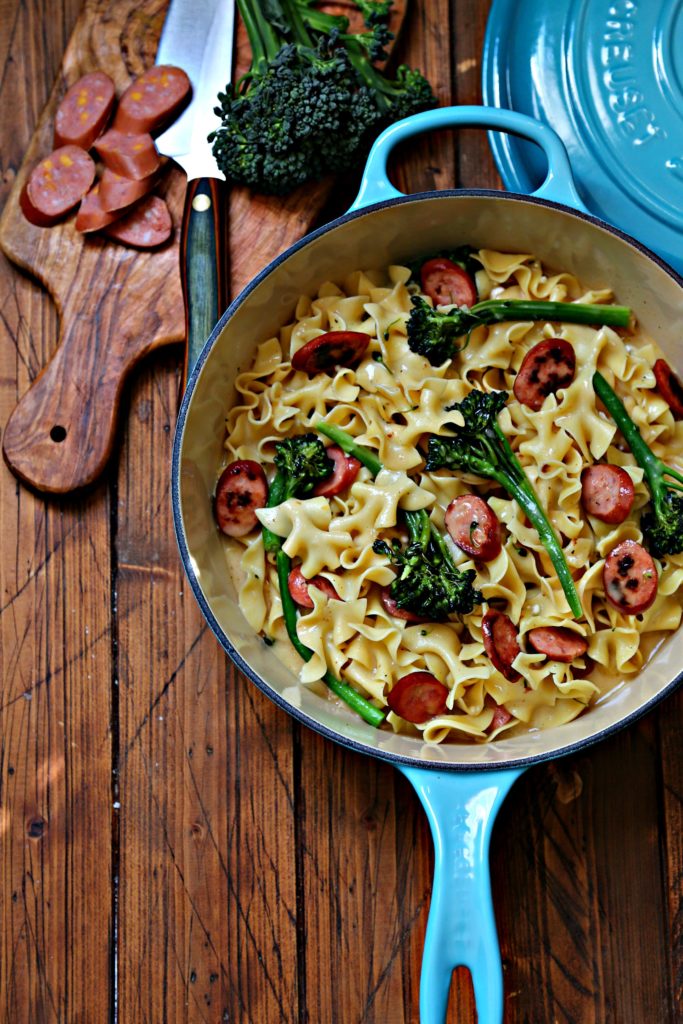 Pasta with sausage and broccoli in blue pot. Cutting board with sliced sausage and knife behind.