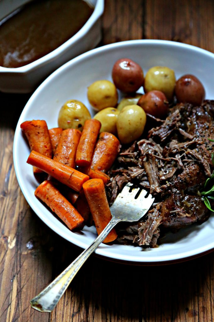 pot roast with carrots and potatoes in white bowl with serving fork. Gravy boat behind.