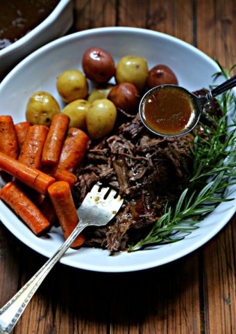 Pot roast, carrots, potatoes in white bowl with spoon of gravy.