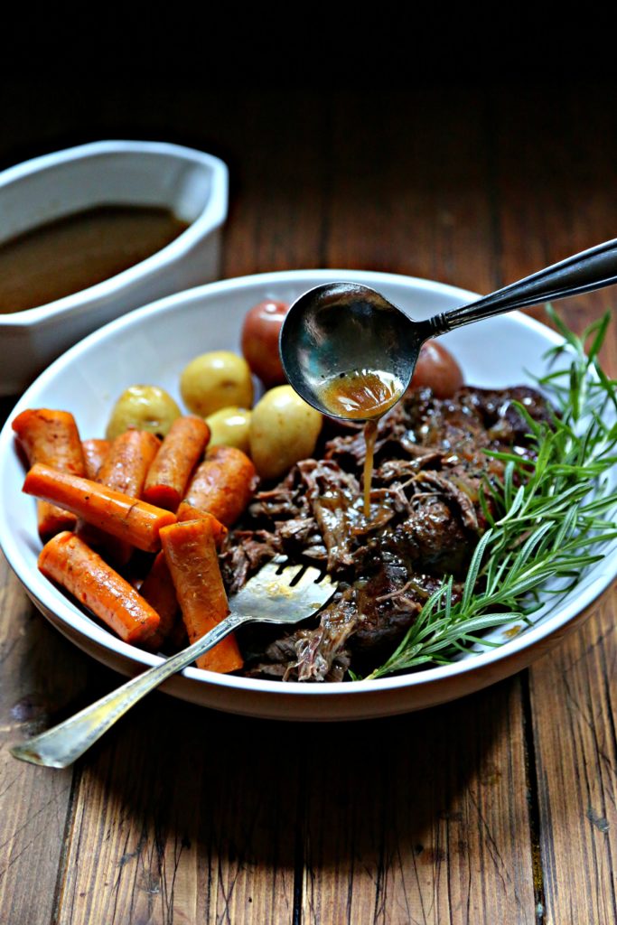 Pot roast, carrots, potatoes in white bowl with spoon of gravy.
