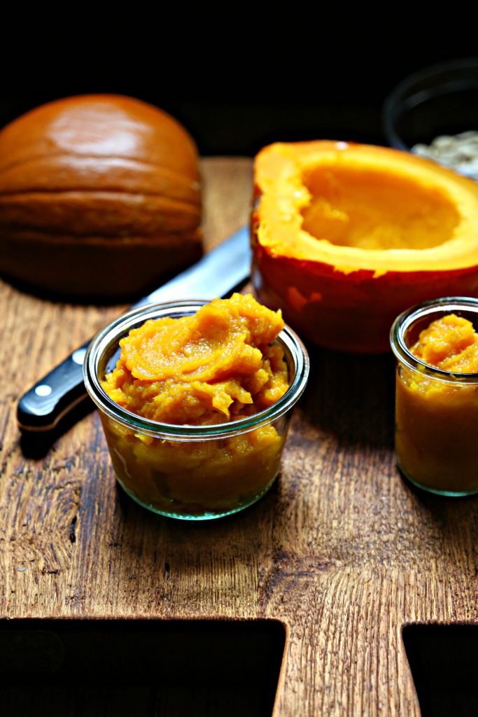 pumpkin puree in small glass jars on cutting board. Knife and pumpkin halves in background.