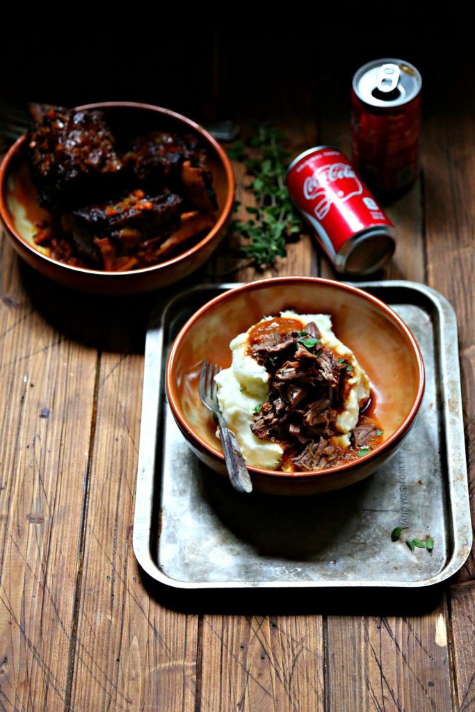 Coca Cola Braised Short Ribs in brown bowl with mashed potatoes. Can of coca cola behind.