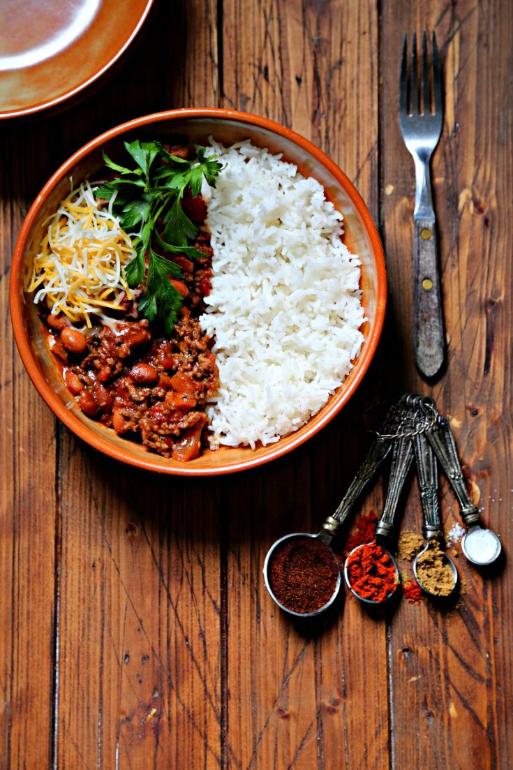 bowl of chili with rice, cheese and parsley. Fork and measuring spoons with spices to side.