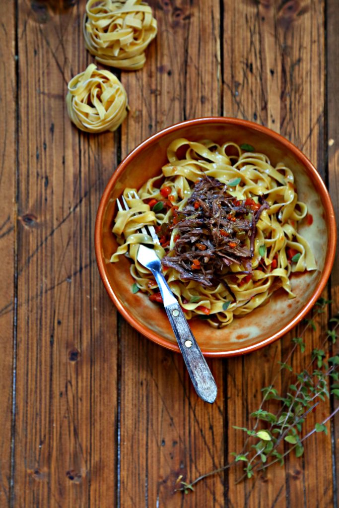 Bowl of pasta with shredded short ribs. Dried pasta nests in background.
