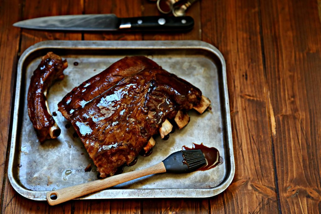 half rack of ribs on baking sheet with silicone brush. Knife and measuring spoons in background.