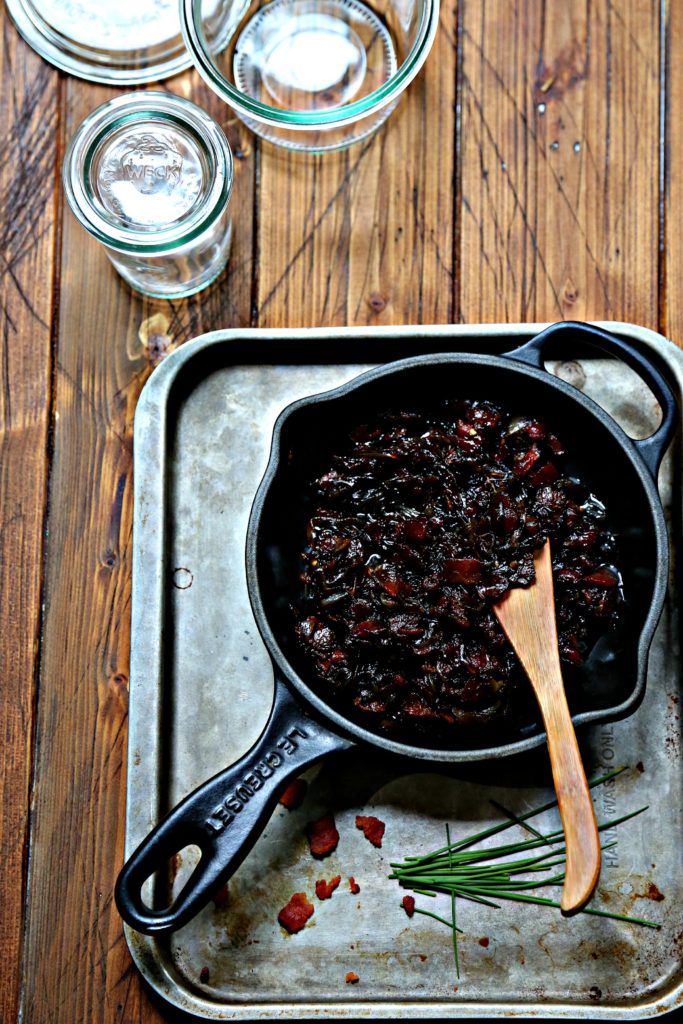 bacon jam in small black skillet on baking sheet with wooden knife. Small glass jars in background. 