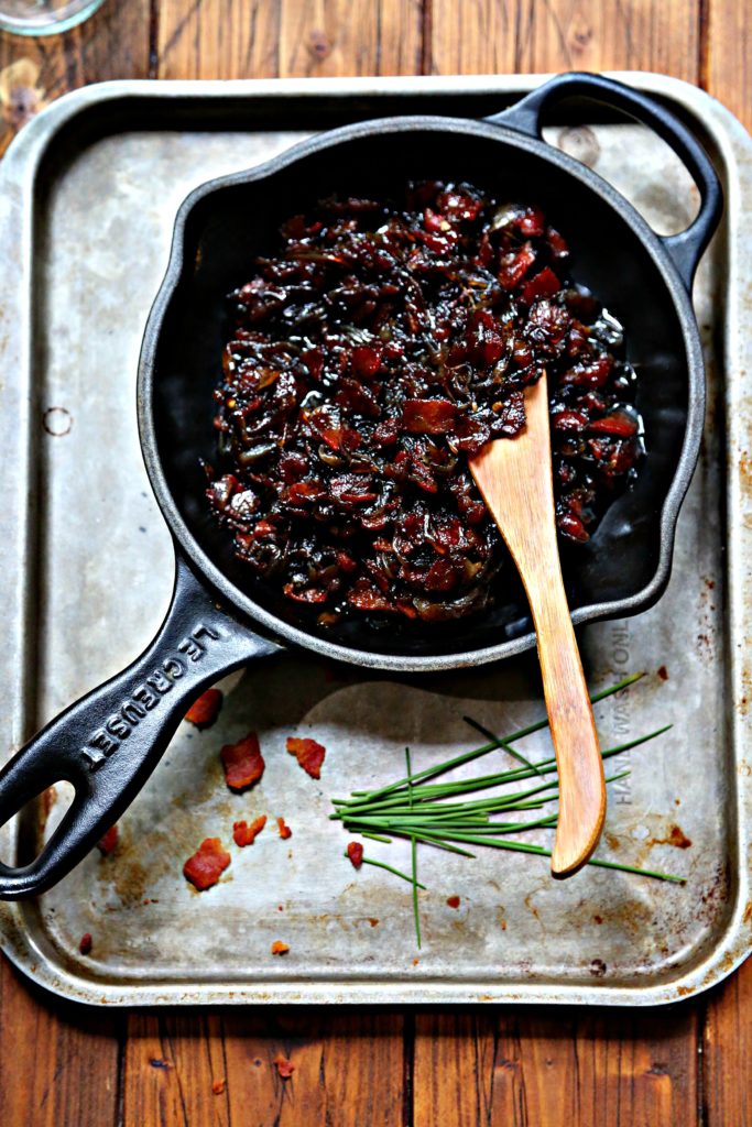 bacon jam in small black skillet on baking sheet. Wooden knife in skillet. Chives and bacon bits on baking sheet.