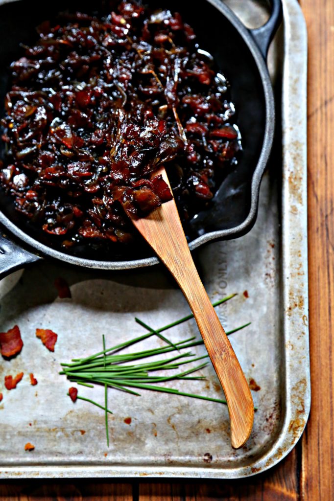 bacon jam in small black skillet on baking sheet with wooden knife. Chives and bacon bits in front.