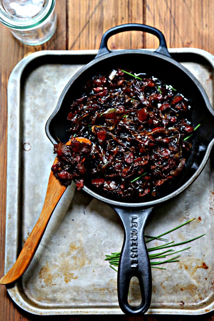 bacon jam in small black skillet on baking sheet. Wooden knife with jam to left. 
