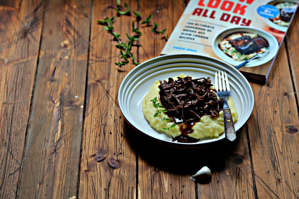 white bowl with mashed potatoes, short ribs and gravy with fork. Cookbook in background. Garlic clove in front of bowl 