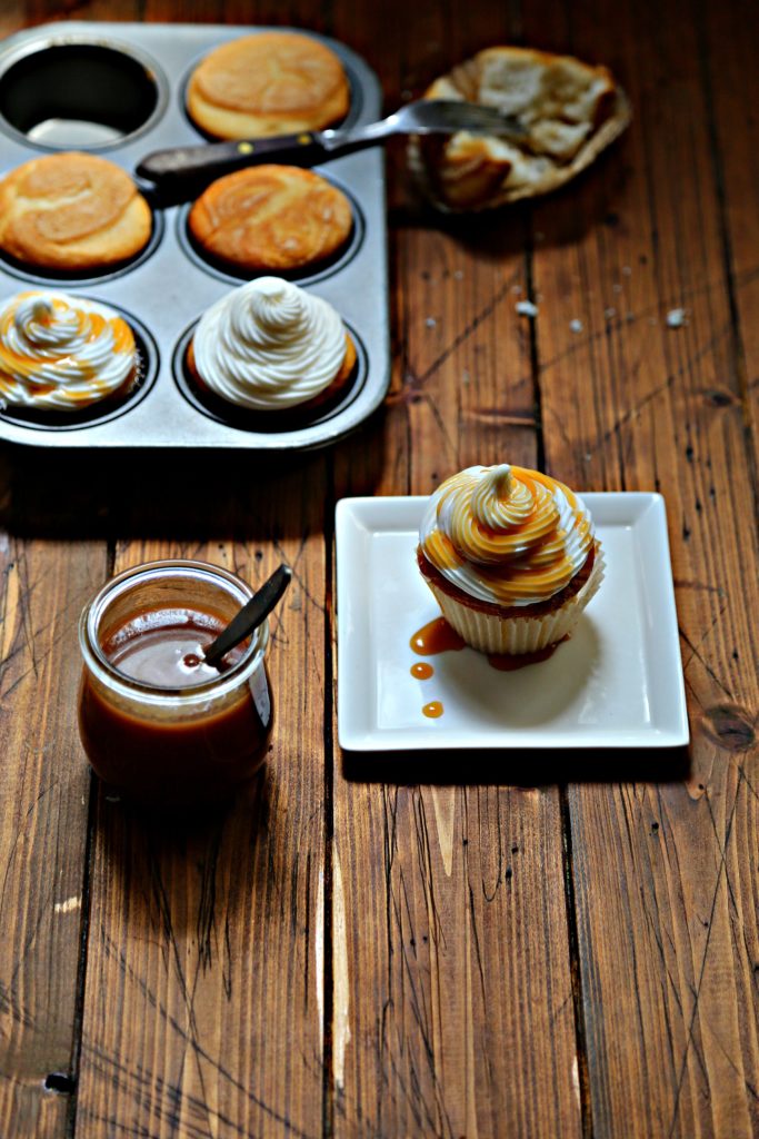 glass jar of caramel sauce with spoon, cupcake on white plate to side, baking tray of cupcakes in background.
