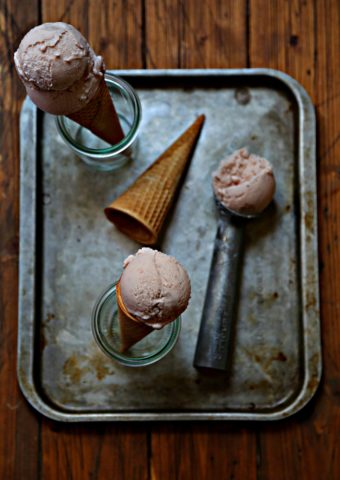 baking sheet with ice cream scooper with scoop of strawberry ice cream. Empty sugar cone laying, 2 ice cream cones standing in small glass jars.