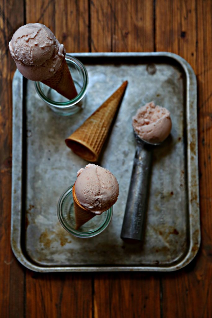 baking sheet with ice cream scooper with scoop of strawberry ice cream. Empty sugar cone laying, 2 ice cream cones standing in small glass jars.