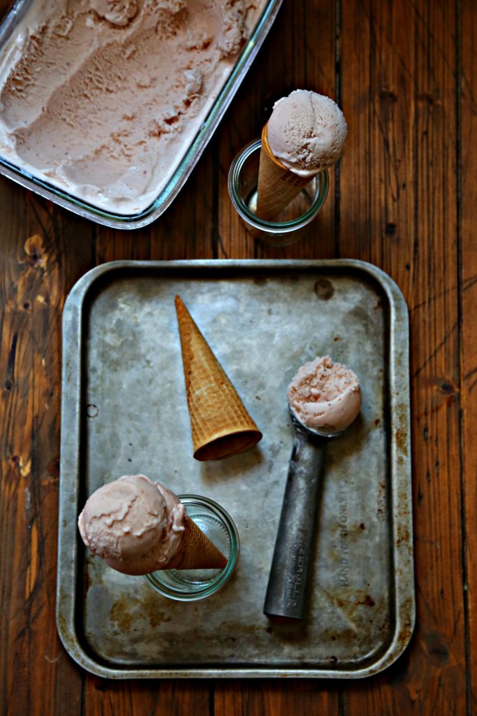baking sheet with ice cream scoop of strawberry ice cream. Sugar cone on its side. Small glass jar holding ice cream cone. Glass container of ice cream in background.