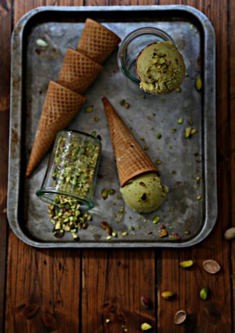 baking sheet with stack of ice cream cones. Small glass of crushed pistachios on its side. Ice cream cone standing in small glass jar.