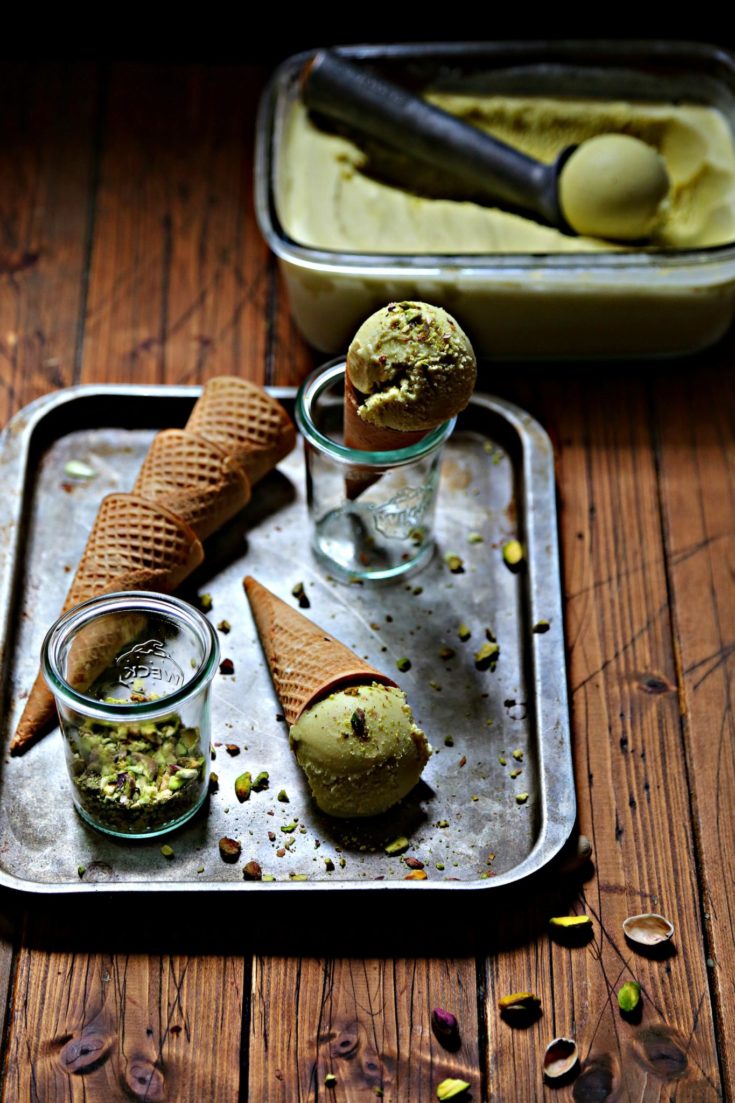 baking sheet with sugar cones, small glass jar of crushed pistachios, pistachio ice cream cone laying flat, pistachio ice cream cone standing in glass jar. Glass container of pistachio ice cream with scoop in background.