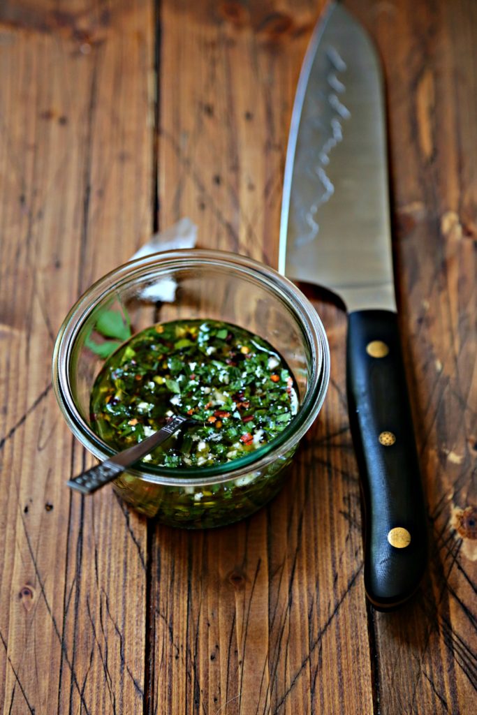 small glass jar with chimichurri sauce and spoon. Knife with black handle to side. 