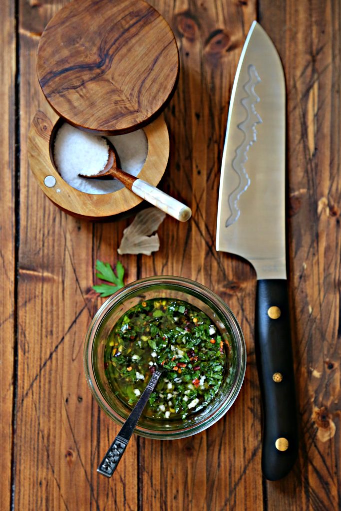 glass jar with chimichurri sauce and spoon. Salt cellar with spoon behind. Knife with black handle to side. 