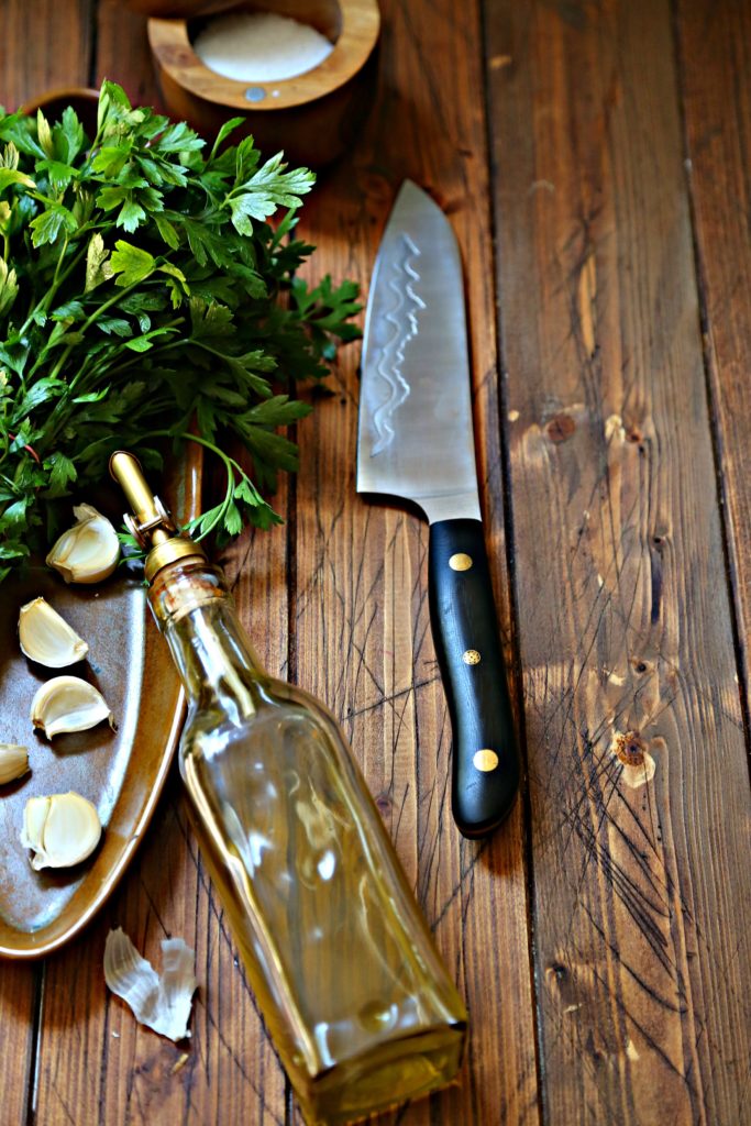 Brown Oblong tray with garlic cloves and bunch of parsley. Olive oil jar leaning against tray. Knife with black handle to side. 