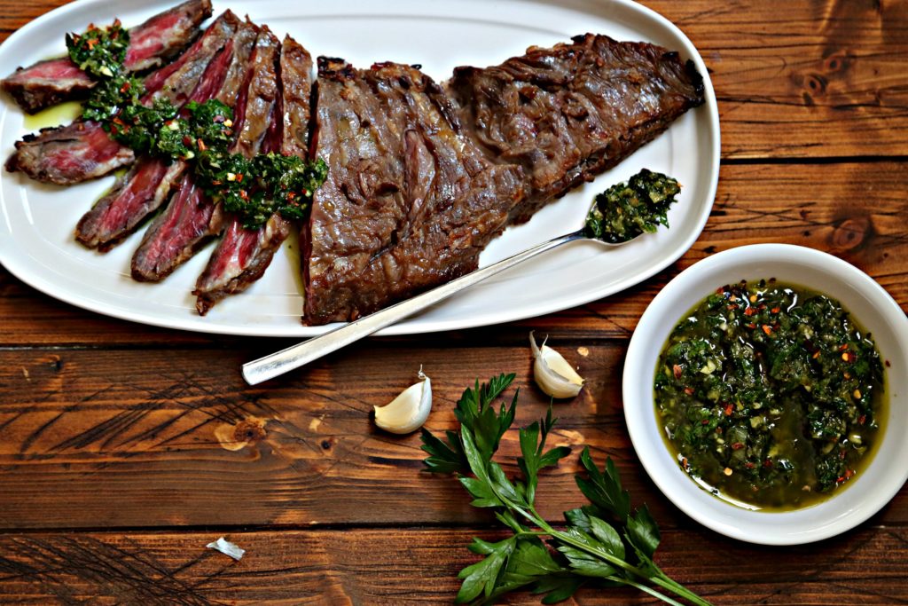 flank steak partially sliced with chimichurri sauce on white plate with fork. Bowl of chimichurri, cloves of garlic and parsley to side.
