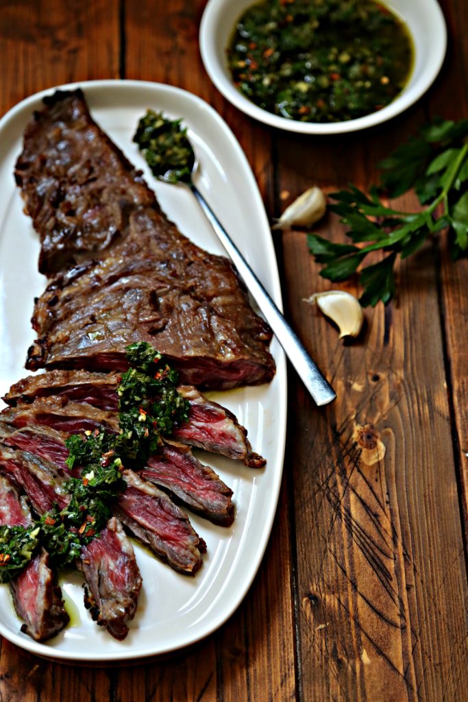 Partially sliced flank steak with chimichurri sauce drizzle over on white plate with spoon. White bowl of chimichurri sauce, cloves of garlic and fresh parsley to side. 