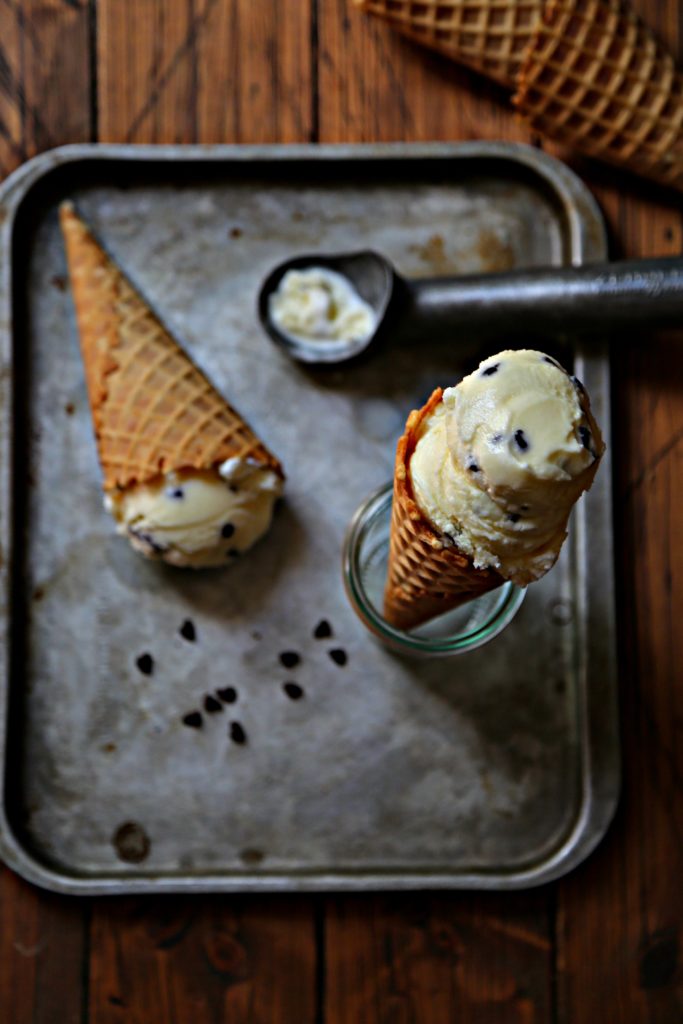 2 mint chocolate chip ice cream cones on baking sheet with ice cream scooper and mini chocolate chips scattered.