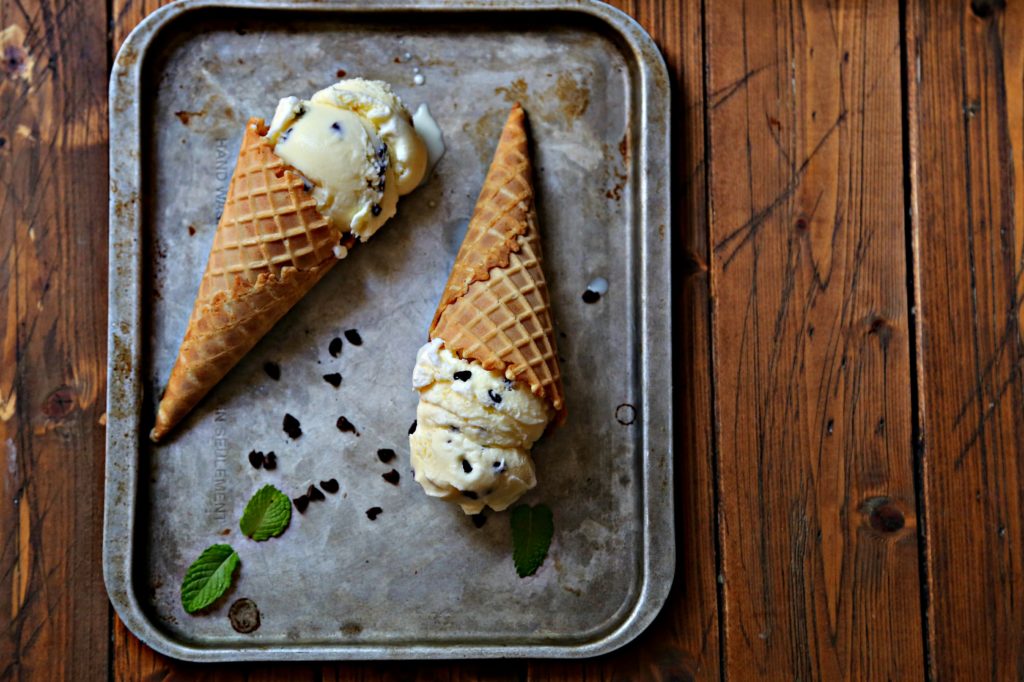 2 mint chocolate chip ice cream cones laying on baking sheet with scattered chocolate chips and mint leaves.