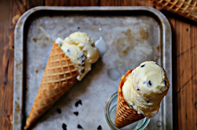 mint chocolate chip ice cream cone laying on baking sheet with chocolate chips scattered. Ice Cream cone standing in glass jar on baking sheet. Container of ice cream and cones in background.